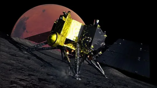 Художественное изображение космического аппарата MMX на темной каменистой поверхности Фобоса. Аппарат выполнен в золотисто-черных тонах, имеет четыре треугольные опоры и темные солнечные панели. На заднем плане виден красный диск Марса. Космический аппарат соберет 10 г лунной пыли с поверхности Фобоса.